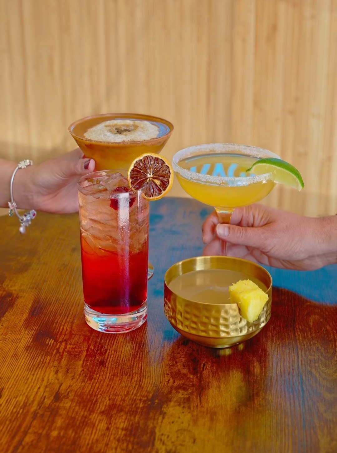 Four colorful drinks on a wooden table with hands reaching for glasses and bowls